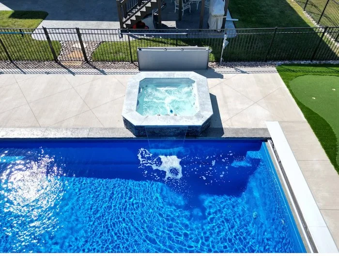 Ground-level detail of a Cardinal fiberglass spillover spa with stone veneer cascading into a Maya Blue Thursday Pools Aspen 16x40.