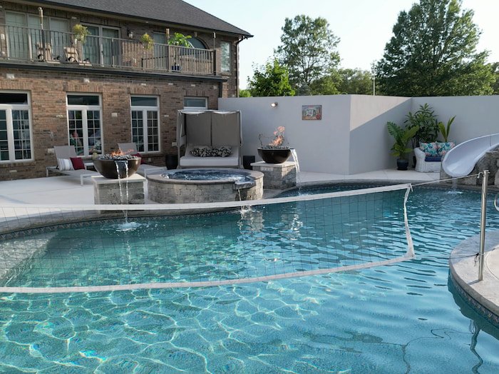 Ground-level view across a 20x42 custom lagoon vinyl liner pool featuring an active volleyball kit, round 12-jet spillover spa with stacked stone surround, and two Grand Effects Legacy fire and water bowls in Beechwood with Tumbled lava rock.