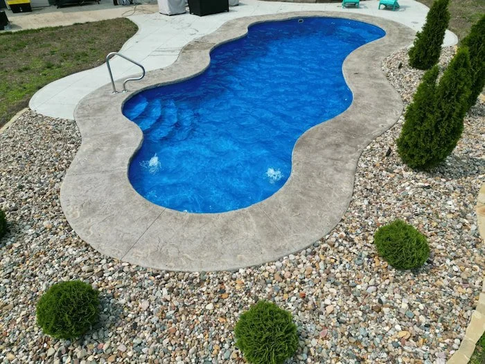 Ground-level view of a Thursday Pools Wellspring 16x40 freeform fiberglass inground pool in Maya Blue with a 3-foot coping band, stamped concrete surround, and rock landscaping.