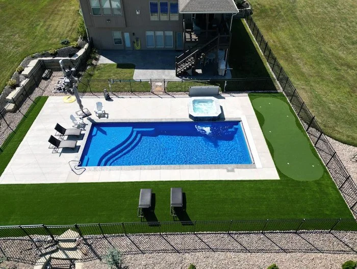 Wide aerial view of a complete Kansas City backyard pool and patio by Tiger Pool and Patio—Thursday Pools Aspen 16x40 in Maya Blue with an automatic safety cover, Cardinal spillover spa, putting green, and turf border on an open lot.