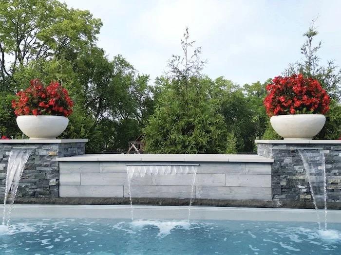 Front-facing view of a 14-foot custom water feature wall with sheer descent waterfall, stone veneer columns, and decorative planters cascading into a Diamond fiberglass inground pool.