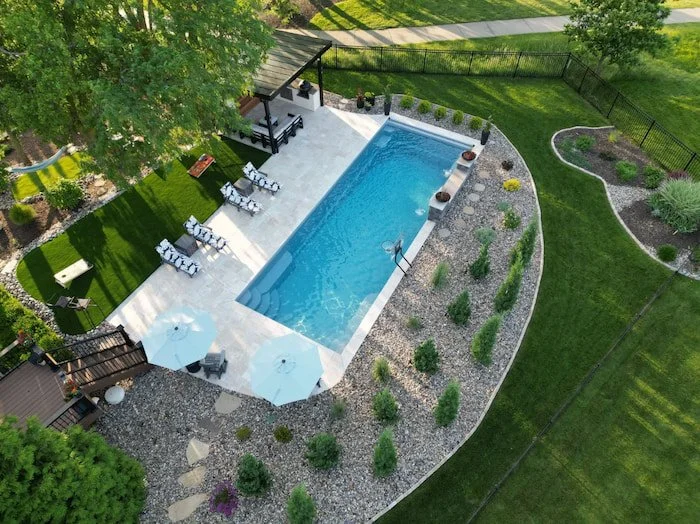 Aerial view of a Thursday Pools Goliath 16x41 fiberglass inground pool installation by Tiger Pool and Patio in Kansas City, featuring a travertine paver patio in a French pattern, basketball goal, water feature wall, and lush professional landscaping