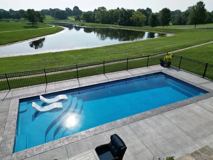 A Thursday Pools Aspen 16x40 fiberglass pool in California Blue with tanning ledge chairs and 16-inch decorative coping, overlooking a scenic waterway, installed by Tiger Pool and Patio in Kansas City.