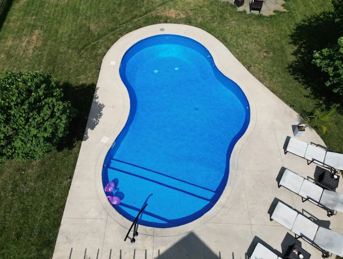 Kansas City fiberglass inground pool design by Tiger Pool and Patio featuring a 20x30 Mountain Lake model with a Merlin Blue Quartz liner, full-length sun deck, black handrail, and surrounding concrete patio with lounge furniture.