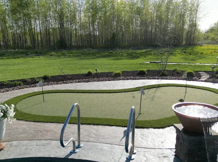 Backyard putting green with synthetic turf and multiple hole flags adjacent to a custom vinyl liner pool with stainless steel entry rails and a copper water bowl feature.
