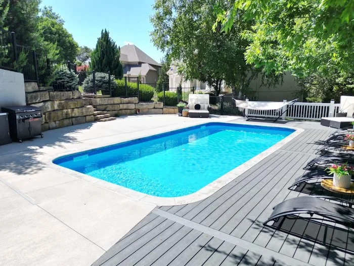 Ground-level view of a Tiger Pool and Patio 14x30 vinyl liner inground pool with a Merlin Blue Quartz liner and corner steps, featuring a gray composite deck surround, natural boulder retaining wall with a stone staircase, outdoor grill station, and 