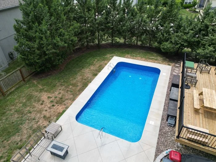 Inground pool installation featuring a 16x32 vinyl liner pool with Jamaica floor pattern, expansive full-length sun deck for maximum lounging, elegant artisan handrail, clean white concrete patio, and seamless integration with natural tree-lined surr