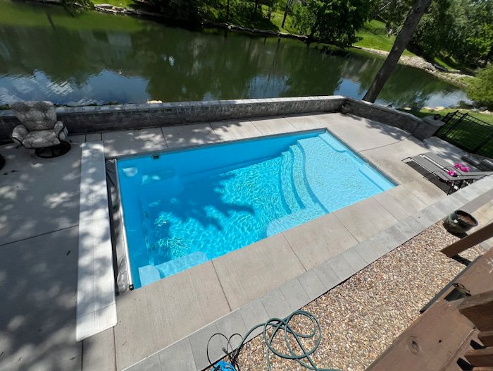Elevated view of a Thursday Pools Aspen 12x25 fiberglass inground pool in Caribbean Sparkle with automatic safety cover, stone retaining walls, and paver coping, set against a stunning Kansas City pond backdrop.