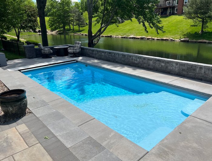 Ground-level view of a Thursday Pools Aspen 12x25 fiberglass inground pool in Caribbean Sparkle with an automatic safety cover, large stone retaining wall, and paver surround overlooking a peaceful Kansas City waterway.