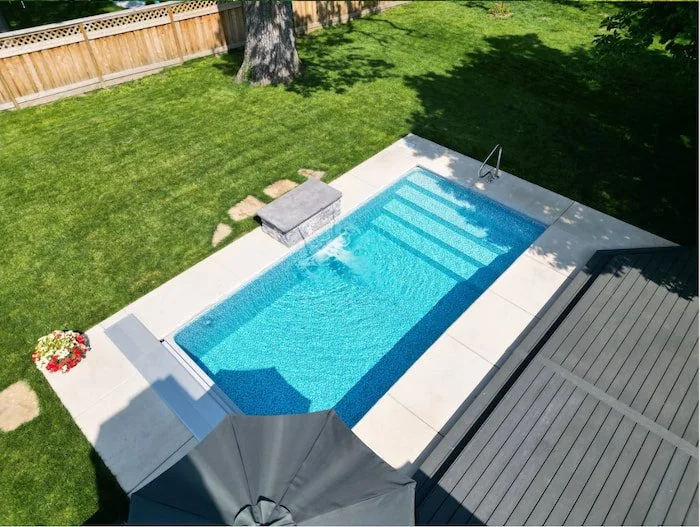 Angled aerial view of a Tiger Pool and Patio 10x20 Recreational personal plunge pool with an Azure Mosaic Borderless liner, active 4-foot stacked stone water feature wall, automatic safety cover, and wide stair entry, installed beside a composite dec