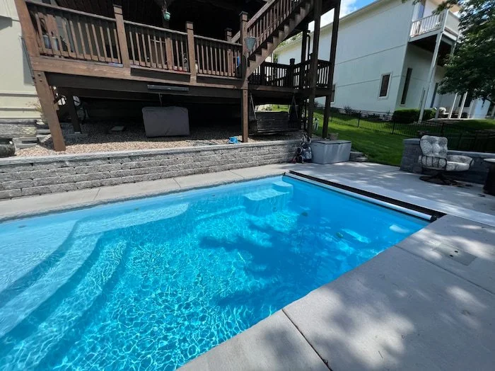 Close view of a Thursday Pools Aspen 12x25 fiberglass inground pool in Caribbean Sparkle with an automatic safety cover and large block retaining wall, tucked beneath a multi-level deck.