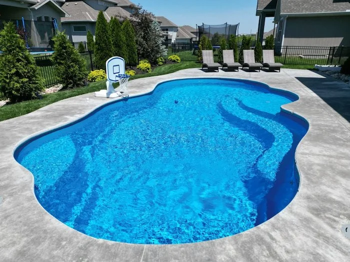 Ground-level view of a Thursday Pools Sun Day 15x30 freeform fiberglass inground pool in California Blue with sun deck bubblers, a basketball goal, and a concrete patio, installed by Tiger Pool and Patio in a landscaped Kansas City backyard.
