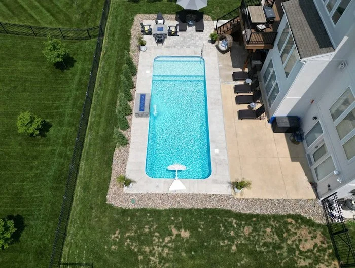 Wide aerial view of a Tiger Pool and Patio 14x30 Serene Tile vinyl liner inground pool with Honey Pearl Floor, showcasing the full-length sun deck, artisan handrail entry, and poolside 4-foot fire and water feature wall.