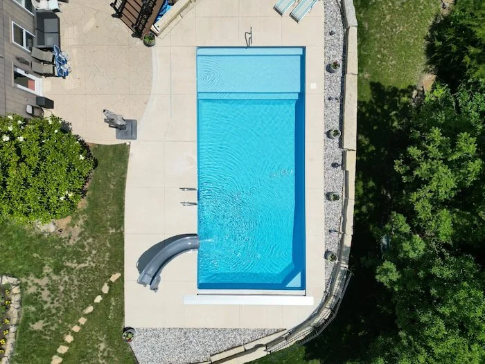 Tiger Pool and Patio Kansas City transforms a sloped backyard with an elegant 18x40 vinyl liner pool featuring a GLI Cayman Mystic Light Blue finish, dramatic Tidal Wave slide, full-length sun deck with corner bench seating, curved natural stone reta