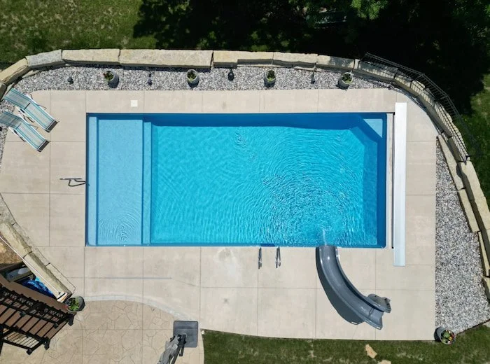 A stunning 18x40 inground swimming pool with a full-length sun deck, corner bench seating, GLI Cayman liner in vibrant Mystic Light Blue, automatic safety cover system, and Tidal Wave slide for family entertainment.