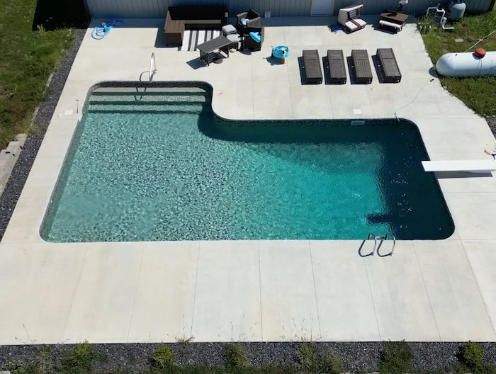 Close aerial view of the GLI Sedona liner and sand gunite color contrast in a Tiger Pool and Patio 16x40x16x26 L-shaped diver inground pool, with full-length steps, a figure four handrail, a diving board, and a deep end ladder visible on a large conc