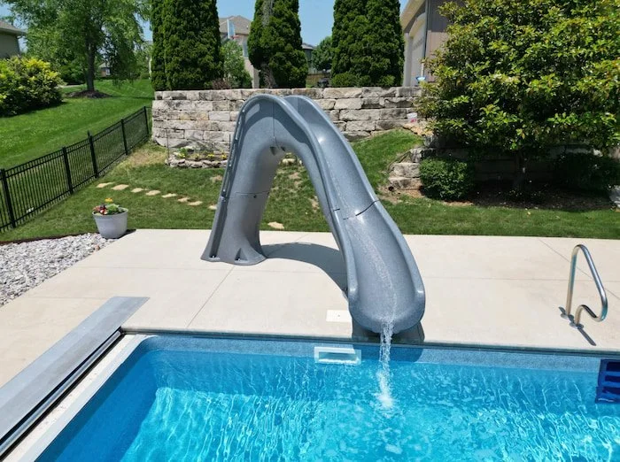 Kansas City pool installation showcasing a premium Tidal Wave slide feature integrated into a 18x40 vinyl liner pool with a GLI Cayman Mystic Light Blue finish, full-length sun deck, natural stone retaining wall, and professional landscaping by Tiger