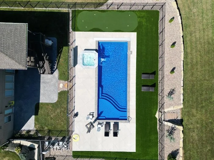 High aerial view of a Thursday Pools Aspen 16x40 fiberglass inground pool in Maya Blue with a Cardinal spillover spa, putting green, and turf border.