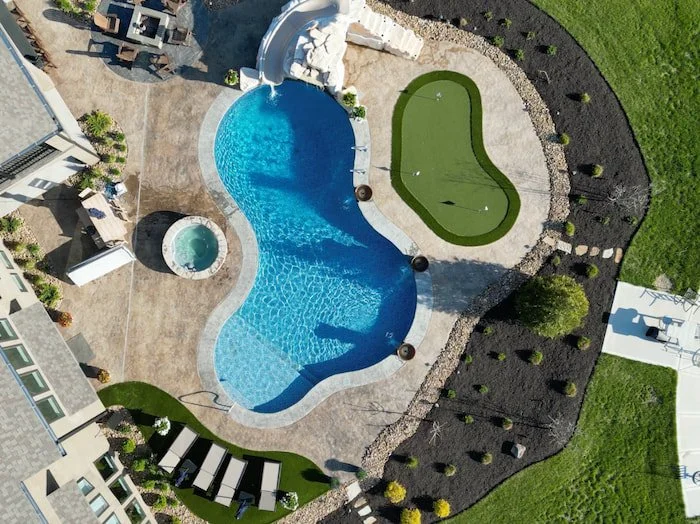 Aerial view of a complete Tiger Pool and Patio backyard installation in Kansas City featuring a 20x42 custom lagoon vinyl liner pool in Opal Bay, stand-alone spa, Rico Rock slide and waterfall, copper water bowls, tanning ledge, synthetic turf puttin