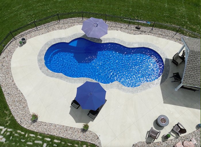 Bird's-eye drone view of the Thursday Pools Sun Day fiberglass inground pool showcasing the signature freeform shape, 16-inch decorative coping band, and expansive concrete pool deck with fire pit seating, a Tiger Pool and Patio installation in Kansa