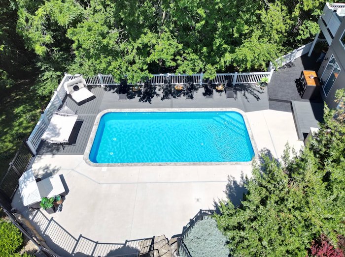 Aerial view of a Tiger Pool and Patio 14x30 Merlin Blue Quartz vinyl liner inground pool with corner steps, surrounded by a gray composite deck with white picket railing, lounge chairs, a hammock stand, and an outdoor bar.