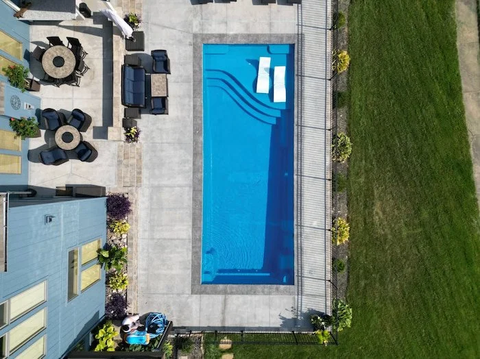 Overhead aerial view of a Thursday Pools Aspen 16x40 fiberglass inground pool in California Blue with tanning ledge chairs, decorative coping, and a concrete patio with fire pit seating area.