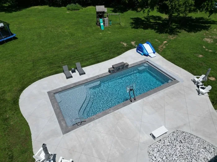 Angled aerial view of a Thursday Pools Aspen 16x40 fiberglass inground pool in Diamond with sheer descent waterfall, salt-friendly basketball goal, automatic safety cover, and concrete patio—a family-friendly inground pool installation by Tiger Pool 