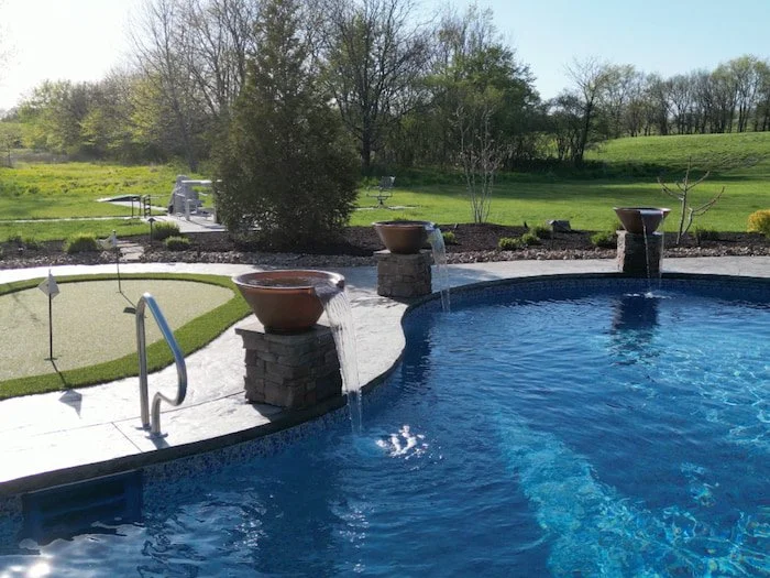Three copper water bowls on stacked stone pedestals cascading into a 20x42 custom lagoon vinyl liner pool in Opal Bay, with a backyard putting green and turf lounge area in the background.