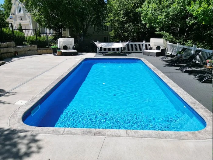 Angled aerial view of a Tiger Pool and Patio 14x30 Merlin Blue Quartz vinyl liner inground pool with corner steps and decorative coping, flanked by a gray composite deck with lounge chairs, hammock stand, and white picket railing, alongside a concret