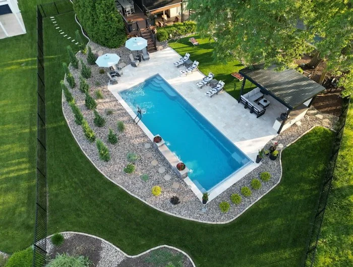 Aerial view of a complete Kansas City backyard pool and patio project by Tiger Pool and Patio—Thursday Pools Goliath 16x41 fiberglass pool in Diamond with a travertine paver surround, pergola lounge area, water feature wall, and lush tiered landscapi