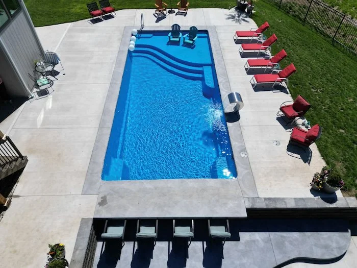 Elevated view of a Thursday Pools Aspen 16x35 fiberglass inground pool in California Blue with a stainless steel waterfall, tanning ledge, and large concrete patio with a sunken outdoor kitchen, installed by Tiger Pool and Patio in Kansas City.