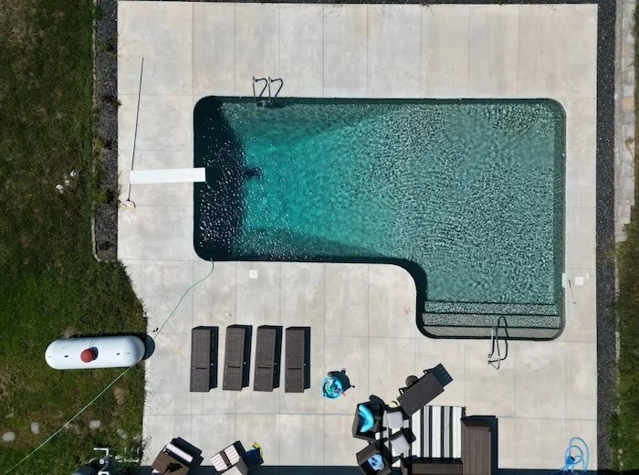 Top-down aerial view of a Tiger Pool and Patio 16x40x16x26 L-shaped diver vinyl liner pool with GLI Sedona liner and sand gunite finish, clearly showing the full-length steps, diving board, figure four handrail, deep end ladder, and large concrete su