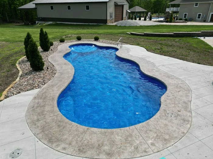 A Thursday Pools Wellspring 16x40 freeform fiberglass inground pool in Maya Blue with stamped concrete surround and evergreen landscaping in a spacious Kansas City backyard.