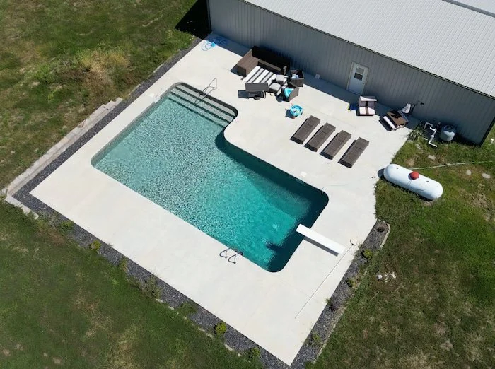 Aerial view of a 16x40x16x26 L-shaped diver pool with GLI Sedona liner and sand gunite shimmering in sunlight, highlighting the figure four handrail, full-length steps, diving board over the deep end, and deep end ladder, set on a concrete patio with
