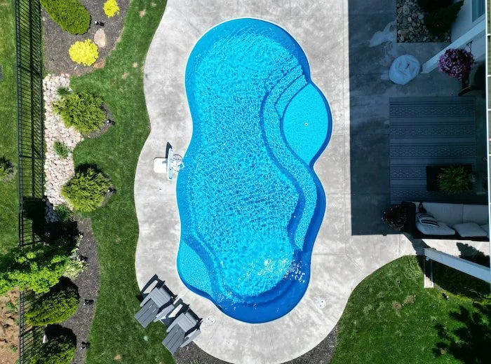 Overhead aerial view of a Thursday Pools Sun Day 15x30 freeform fiberglass inground pool in California Blue with sun deck bubblers, a basketball goal, and a concrete surround.