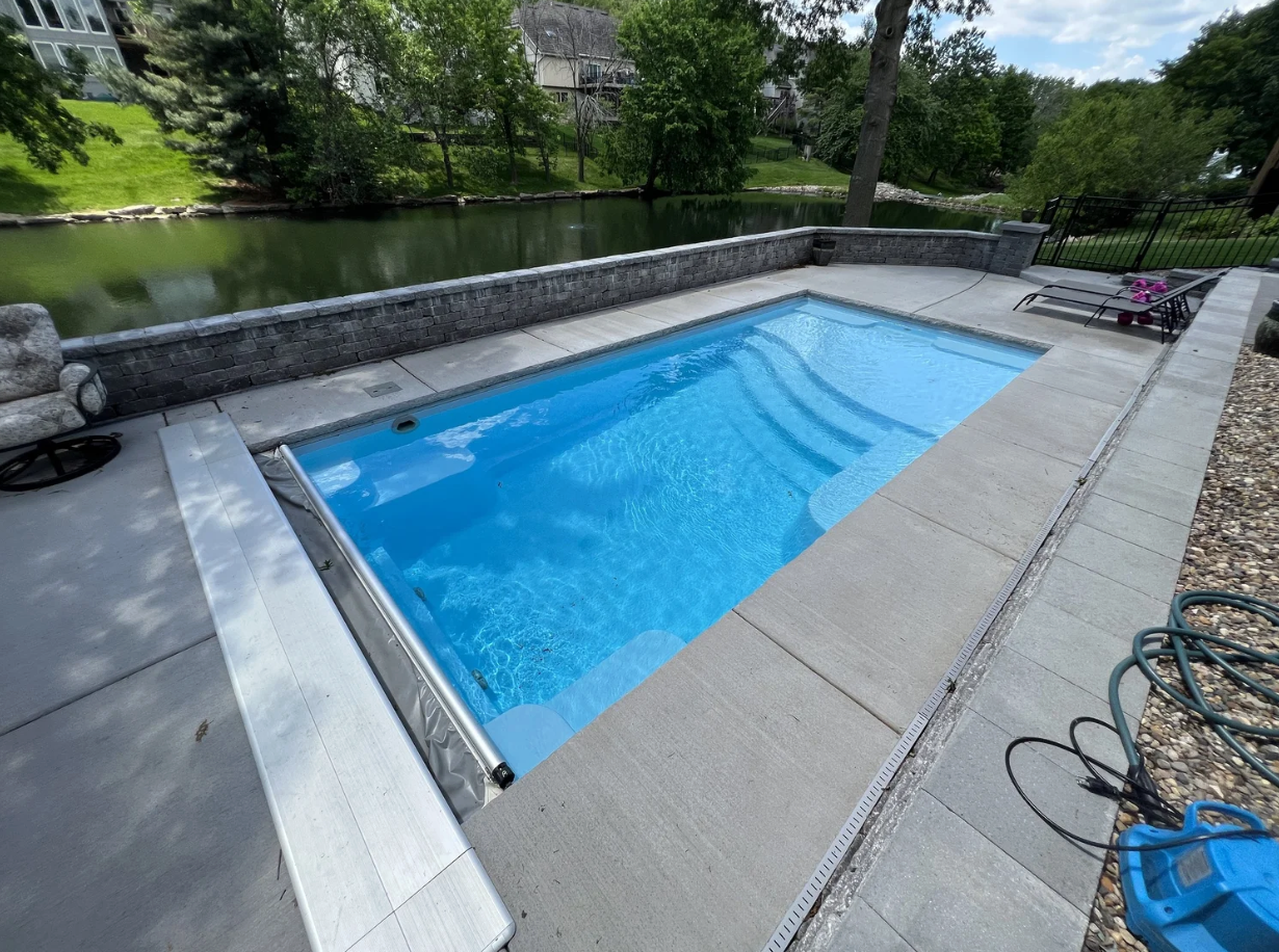  Thursday Pools Aspen 12x25 fiberglass inground pool in Caribbean Sparkle with an automatic safety cover and stone retaining wall.