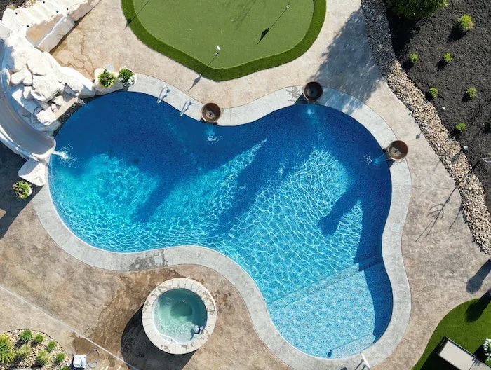 Aerial drone view of a Tiger Pool and Patio 20x42 custom lagoon inground pool in Opal Bay vinyl liner, featuring a tanning ledge, stand-alone spa, three copper water bowls, a Rico Rock slide and waterfall, circular putting green, and stamped concrete
