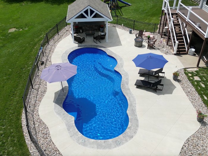 Aerial view of a complete Kansas City backyard transformation by Tiger Pool and Patio, featuring the Thursday Pools Sun Day 16x39 fiberglass pool in Maya color with sun deck bubblers, a custom pool house pavilion, fire pit lounge area, and river rock