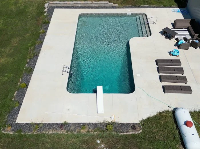 Angled aerial view of a Tiger Pool and Patio 16x40x16x26 L-shaped diver vinyl liner pool with GLI Sedona liner and sand gunite, featuring a diving board, full-length steps, figure four handrail, and deep end ladder on a large concrete pool deck in a 