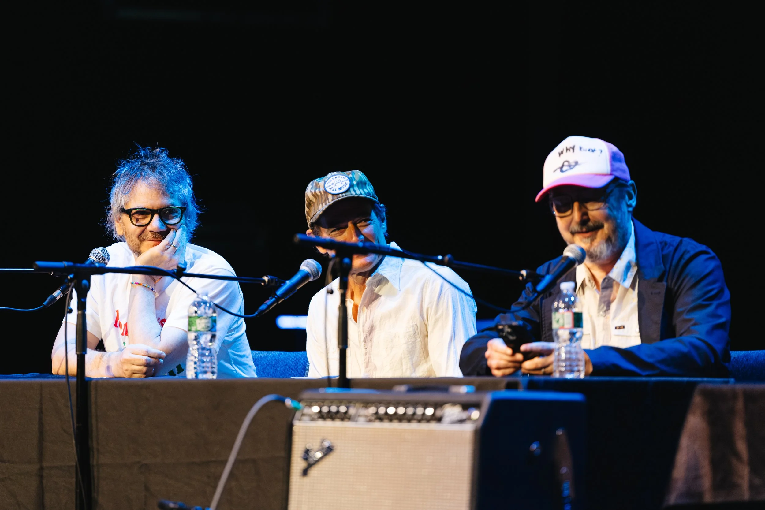 Three men sitting at a table with microphones, water bottles, and papers, engaging in a discussion on stage with a black background.
