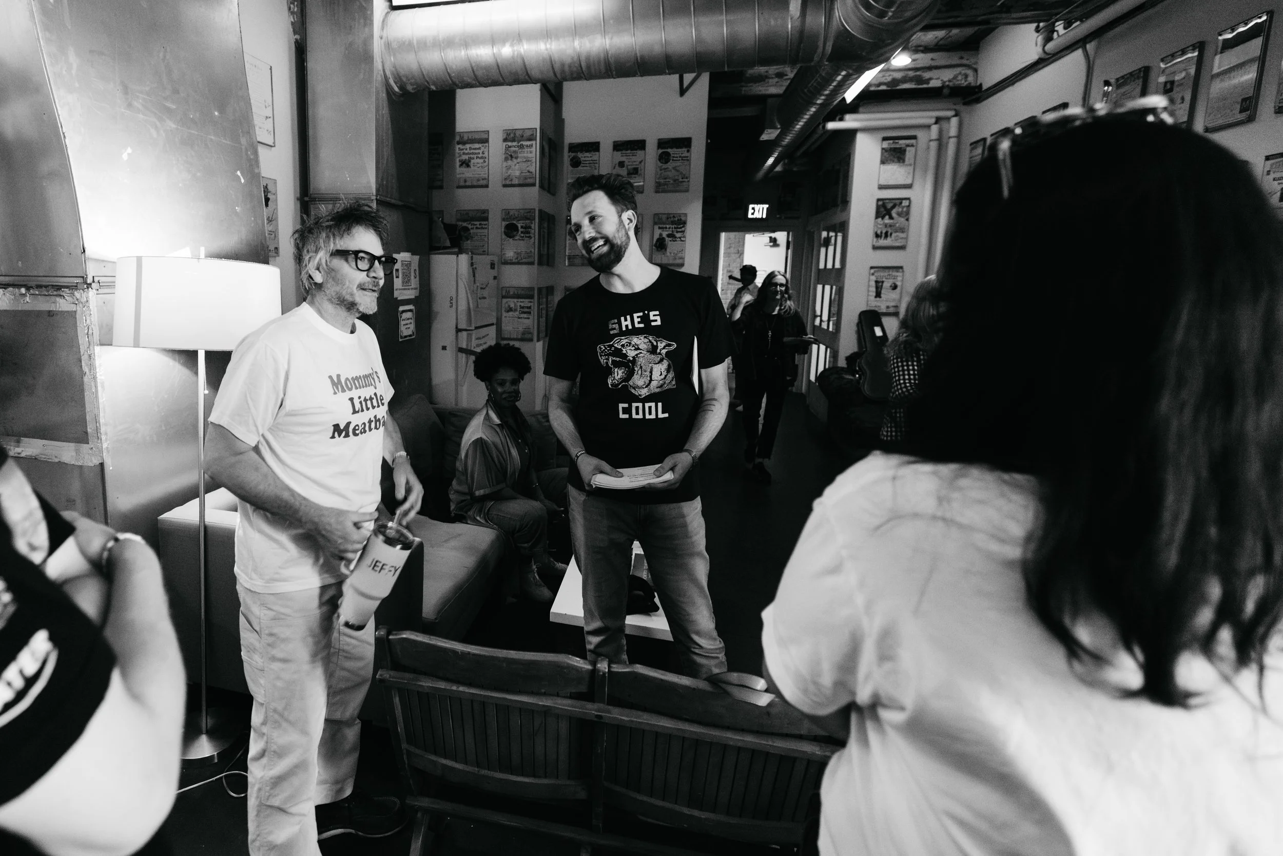 Jeff Tweedy and Jordan Klepper backstage. People gathered indoors, smiling and engaging in conversation, some sitting on chairs and others standing, with posters on the wall and a lamp on a side table.