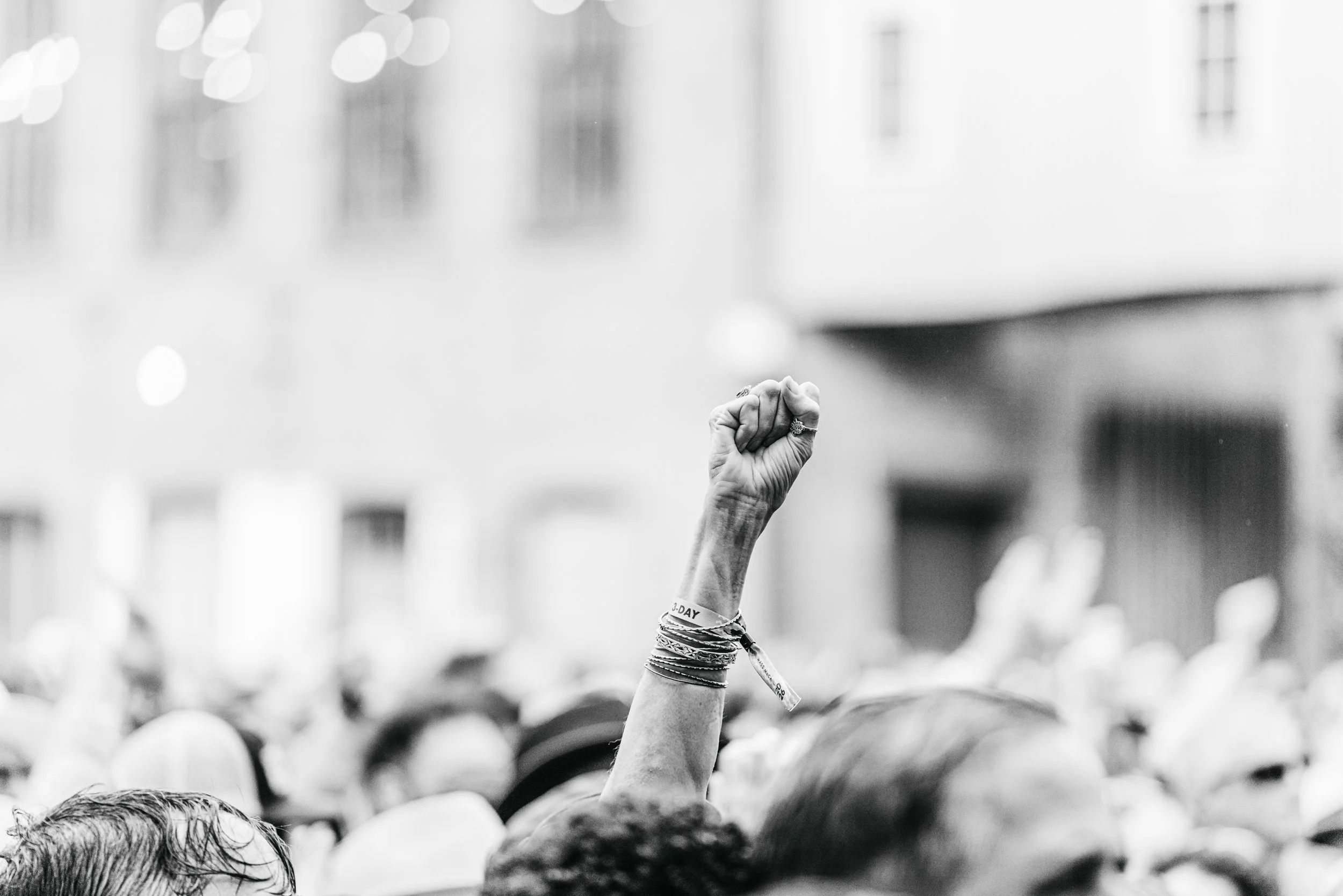 A raised fist of an individual in a crowd at a protest or rally, wearing multiple bracelets and rings, with a blurred background of people and buildings.