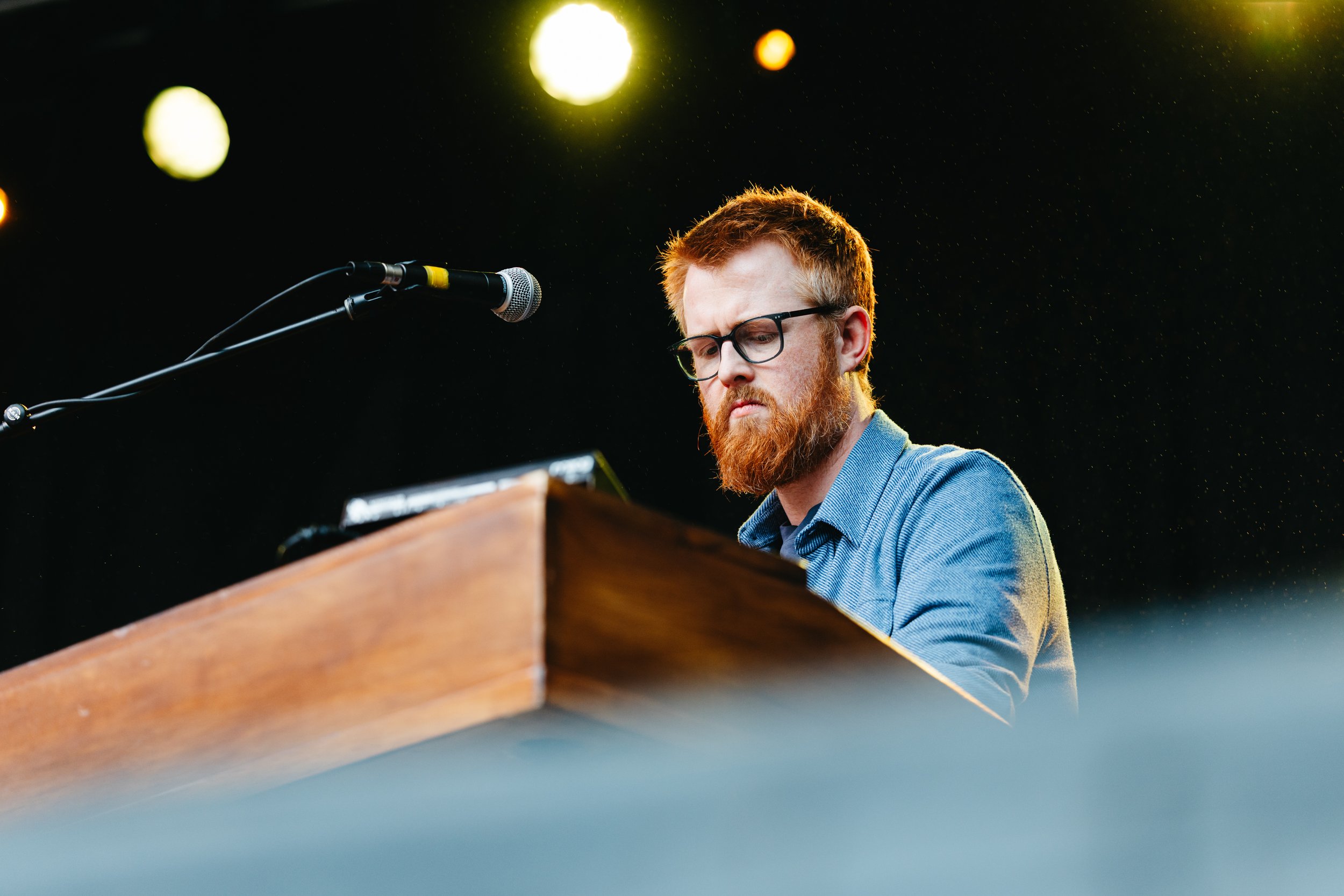 A man with red hair, beard, and glasses playing a piano on stage under bright lights.