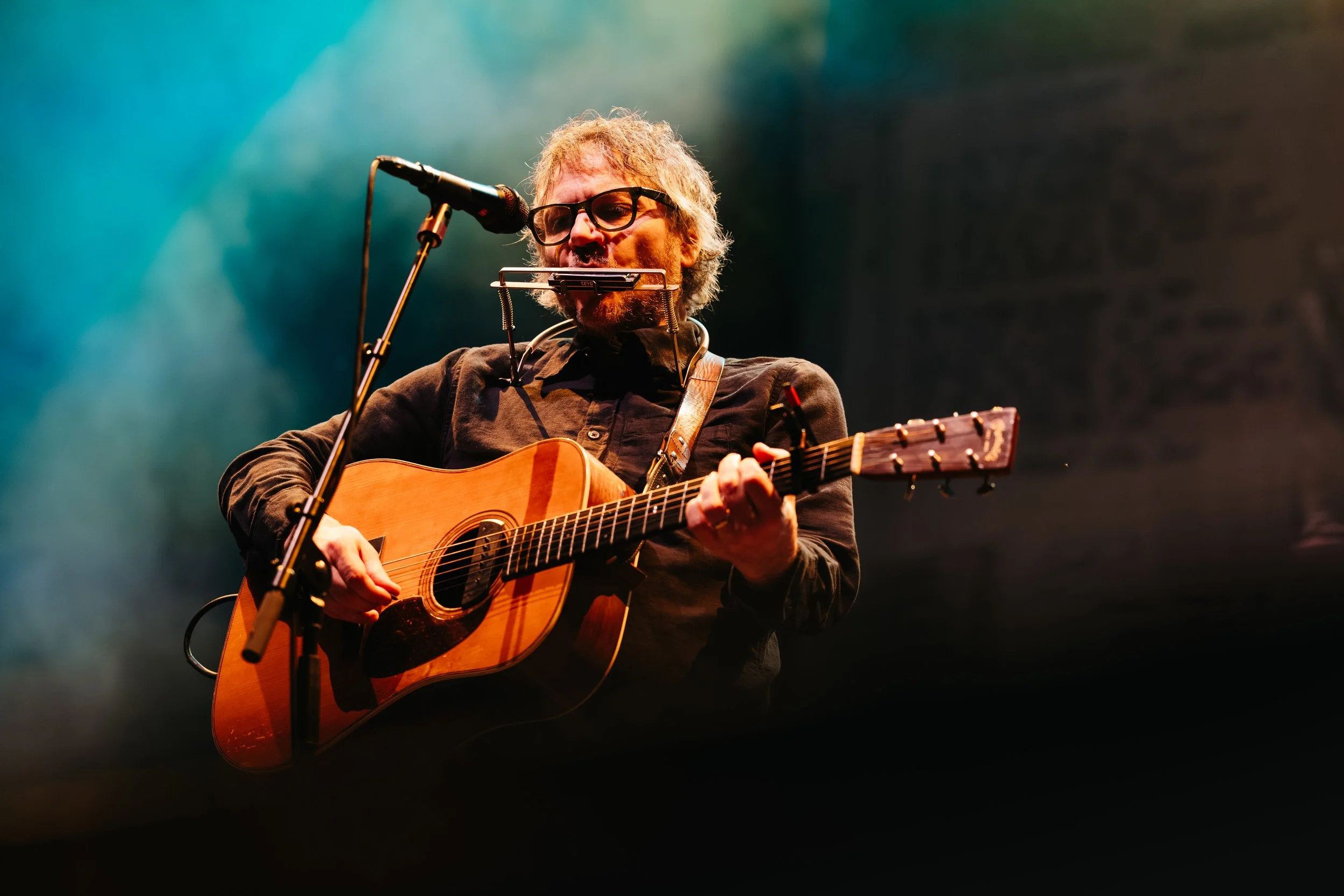 A musician with glasses and curly hair is playing an acoustic guitar on stage, with a harmonica holder around his neck, under colorful stage lighting.