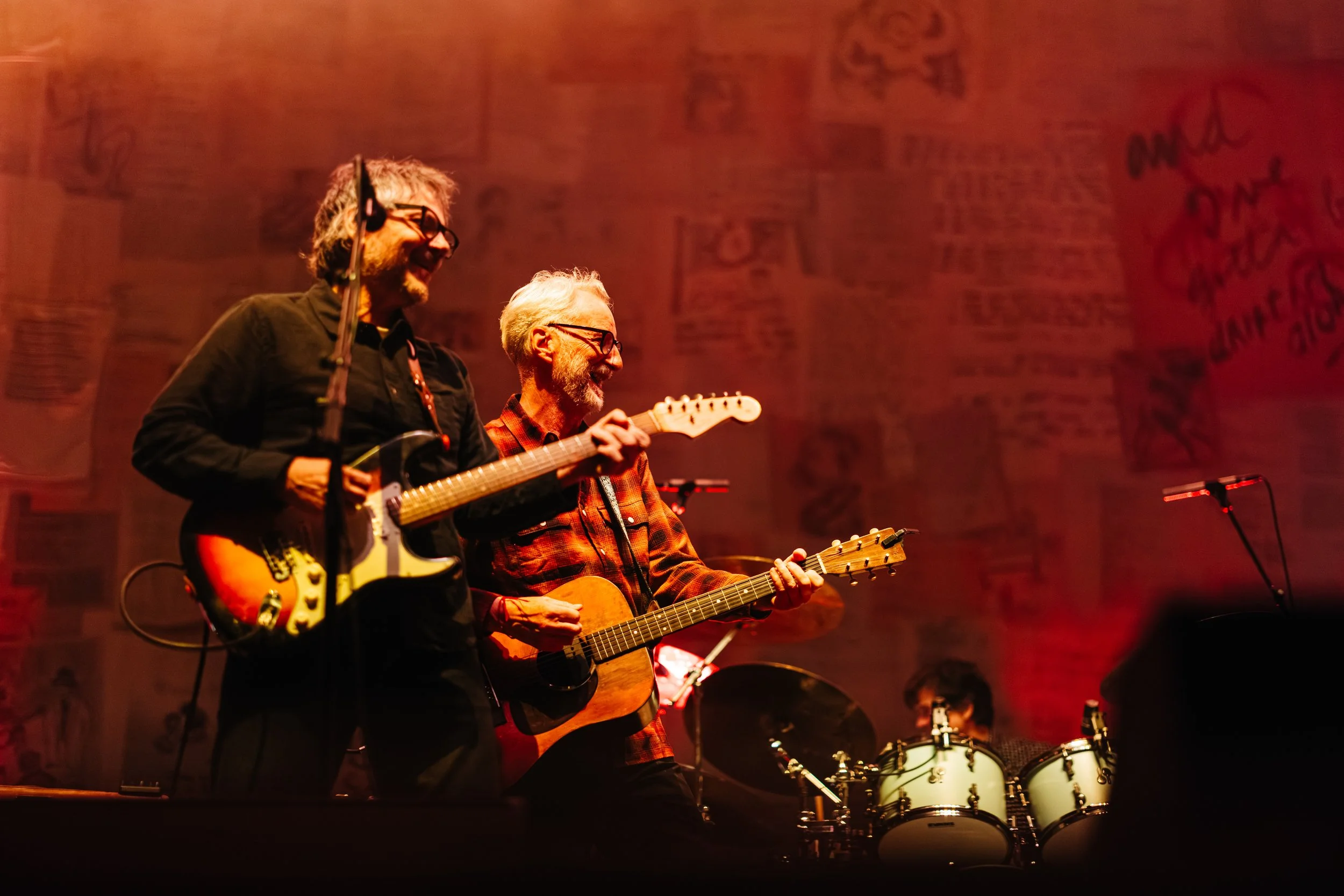 Two musicians performing on stage: one playing electric guitar and the other playing acoustic guitar, both smiling, with drum set in the background, stage illuminated with warm red lighting.
