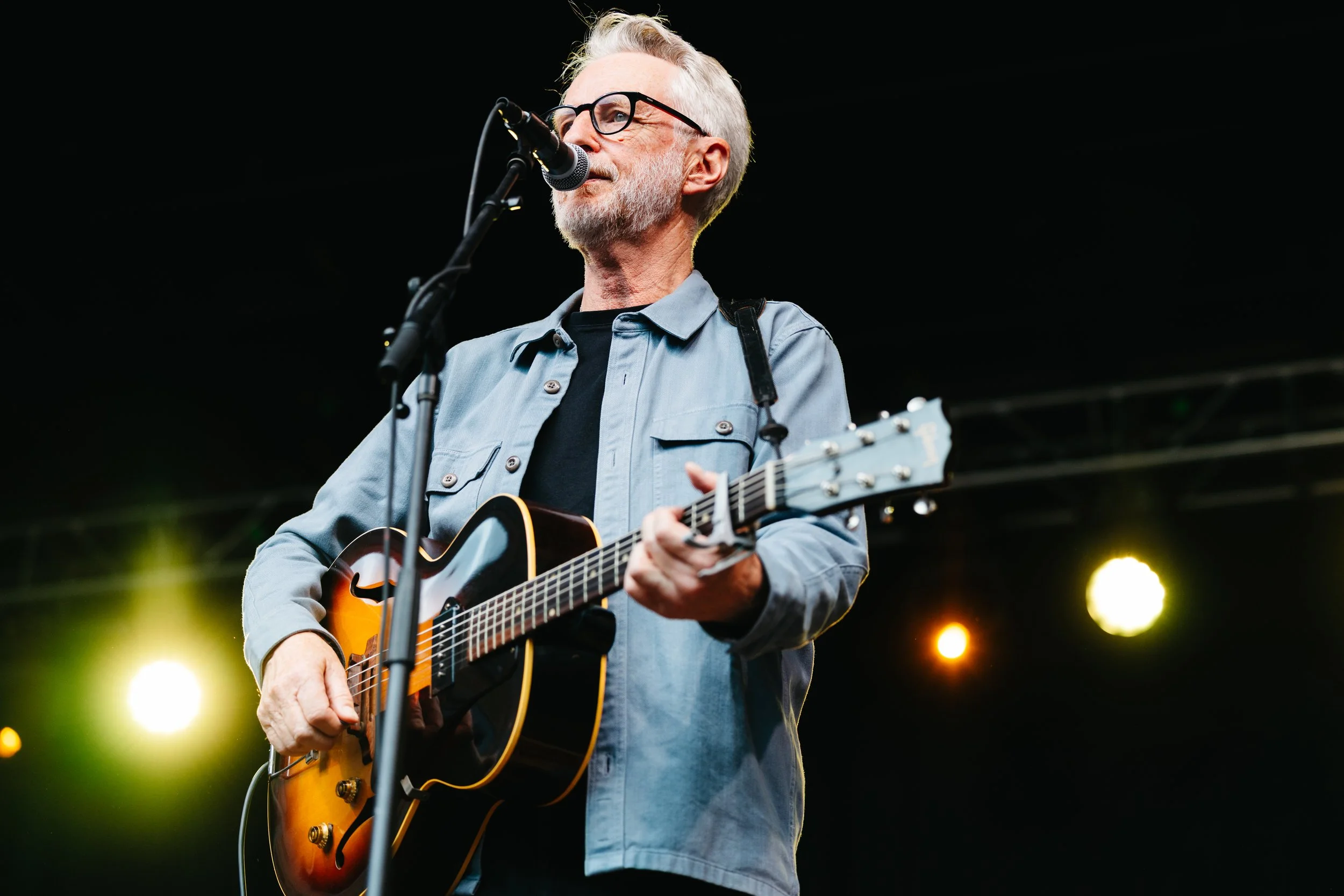 An older man with white hair, beard, and glasses, wearing a denim shirt and black t-shirt, is playing an acoustic guitar while singing into a microphone on stage with colorful lights in the background.