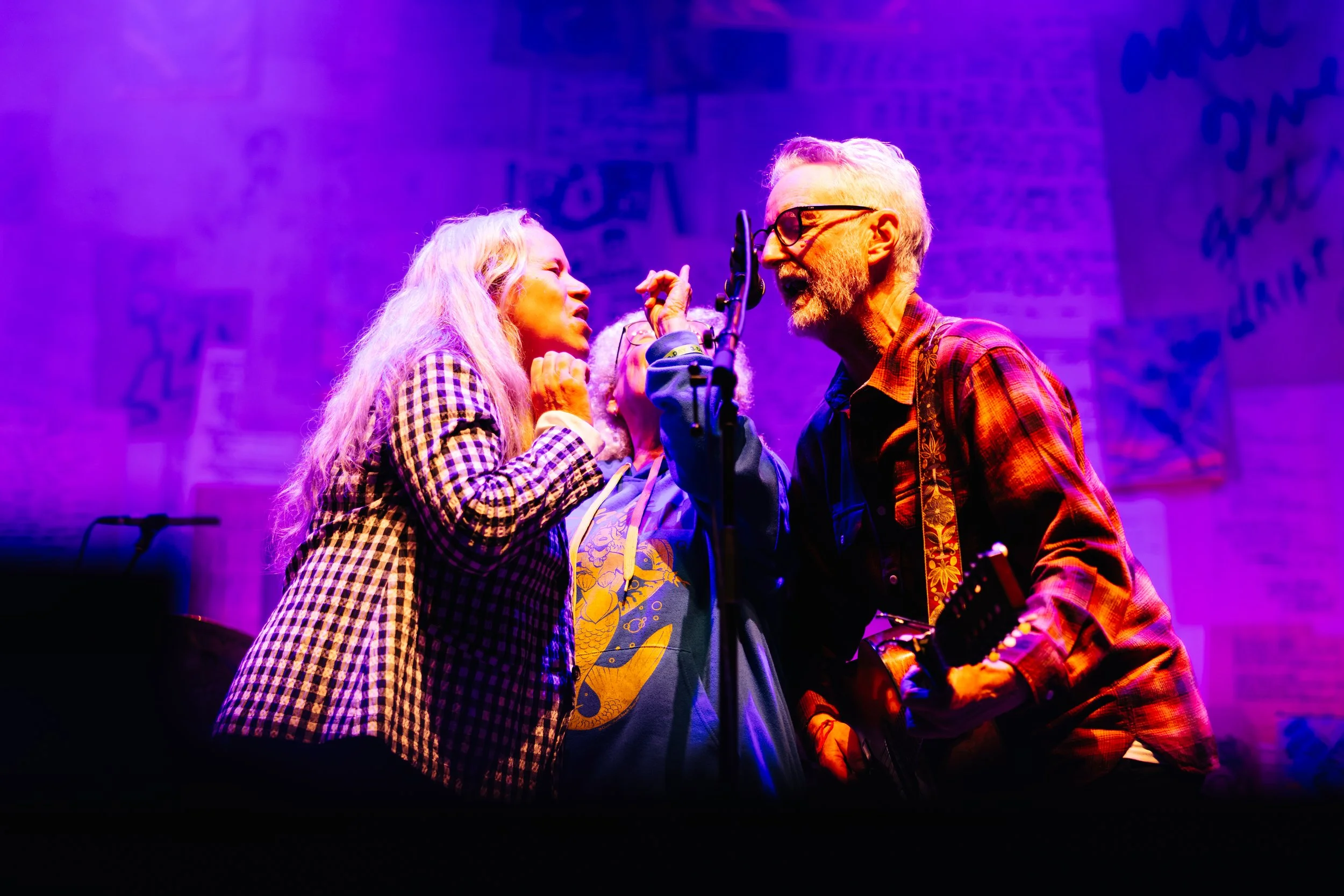 Two musicians, a woman and a man, are singing into a microphone on stage with purple and orange lighting, during a live performance.