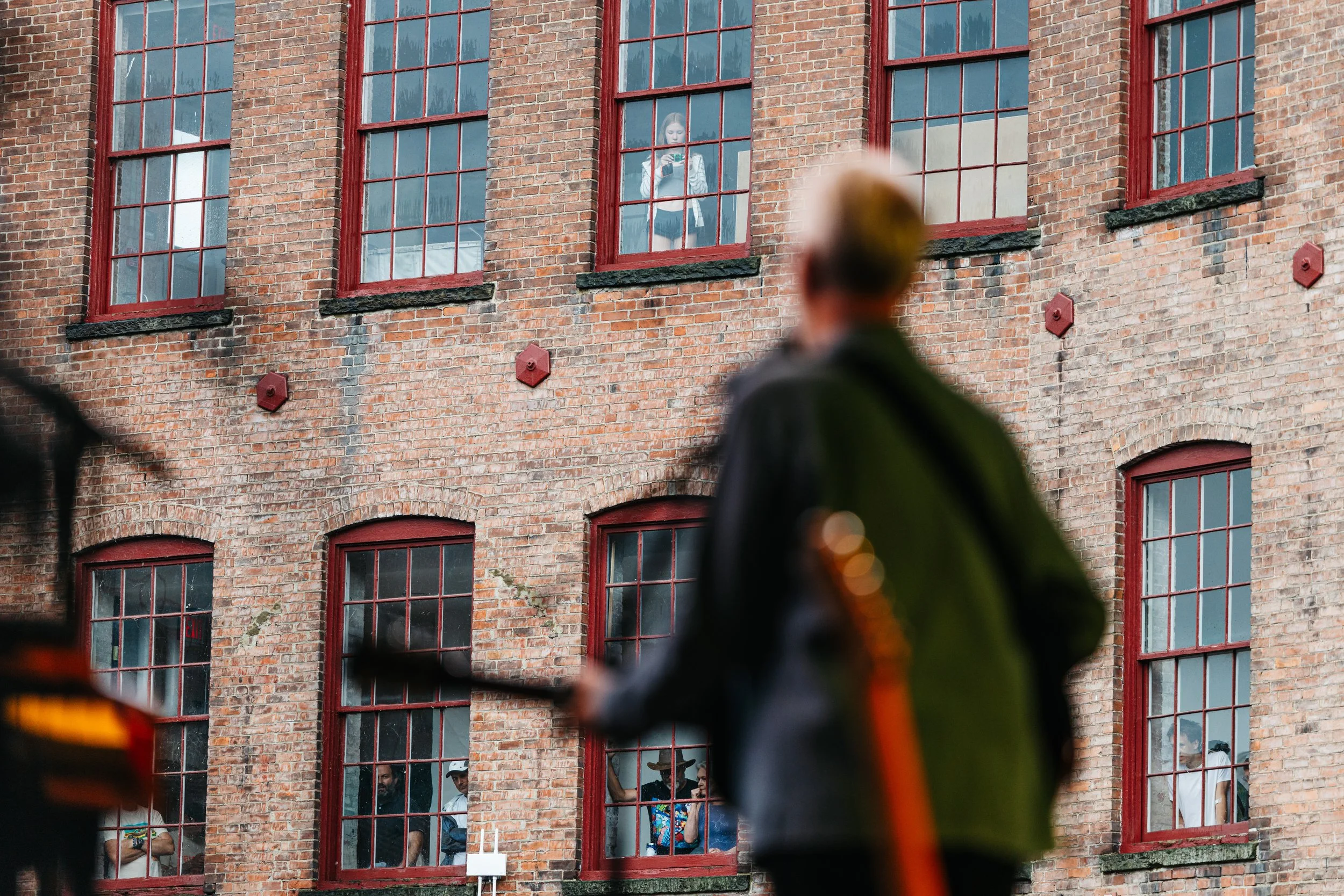 A woman with a striped shirt talking on her phone and a man looking out of a window in a brick building, seen through blurred construction helmets in the foreground.