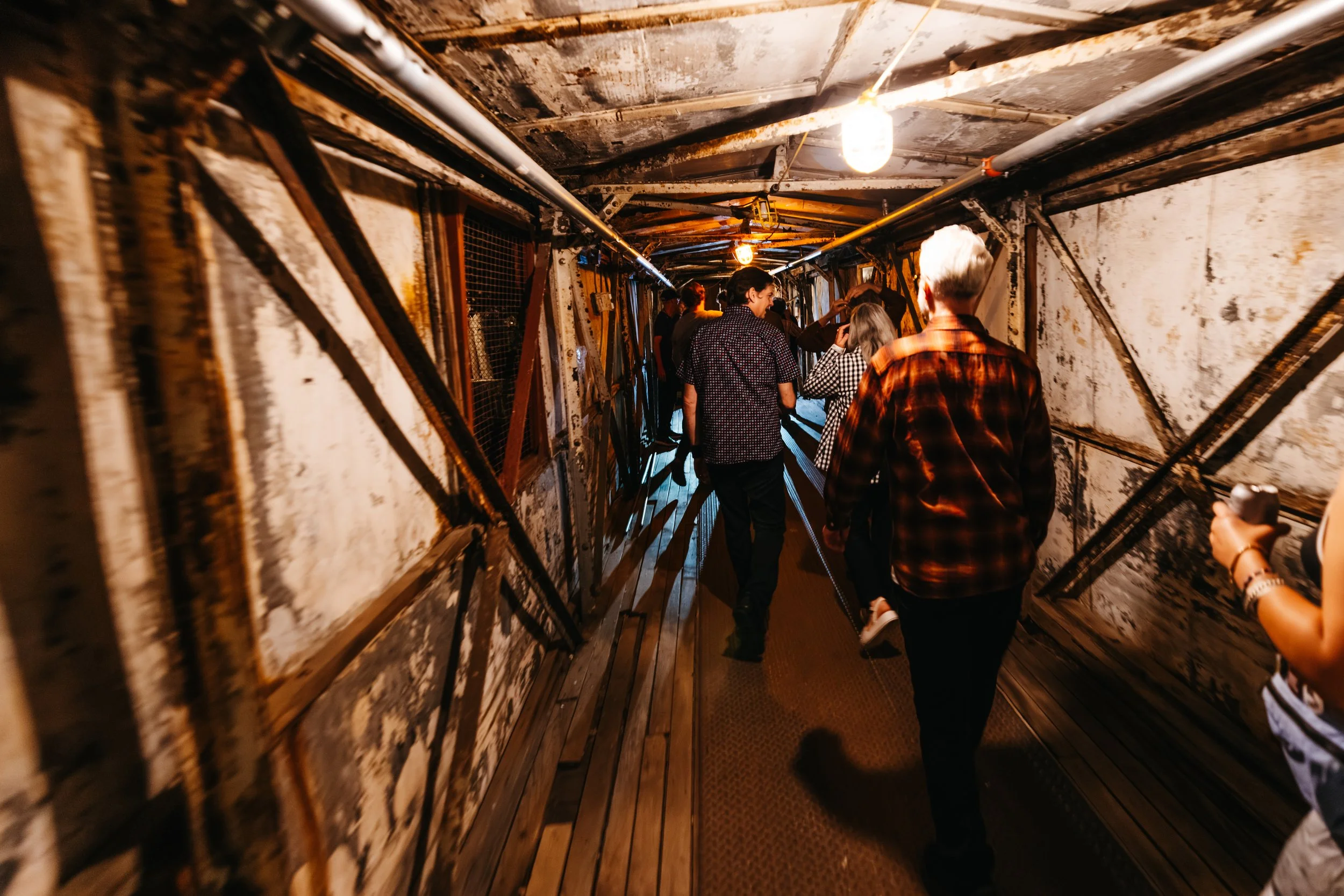 People walking through a narrow, rusted, industrial-looking tunnel with exposed pipes and graffiti on the walls.