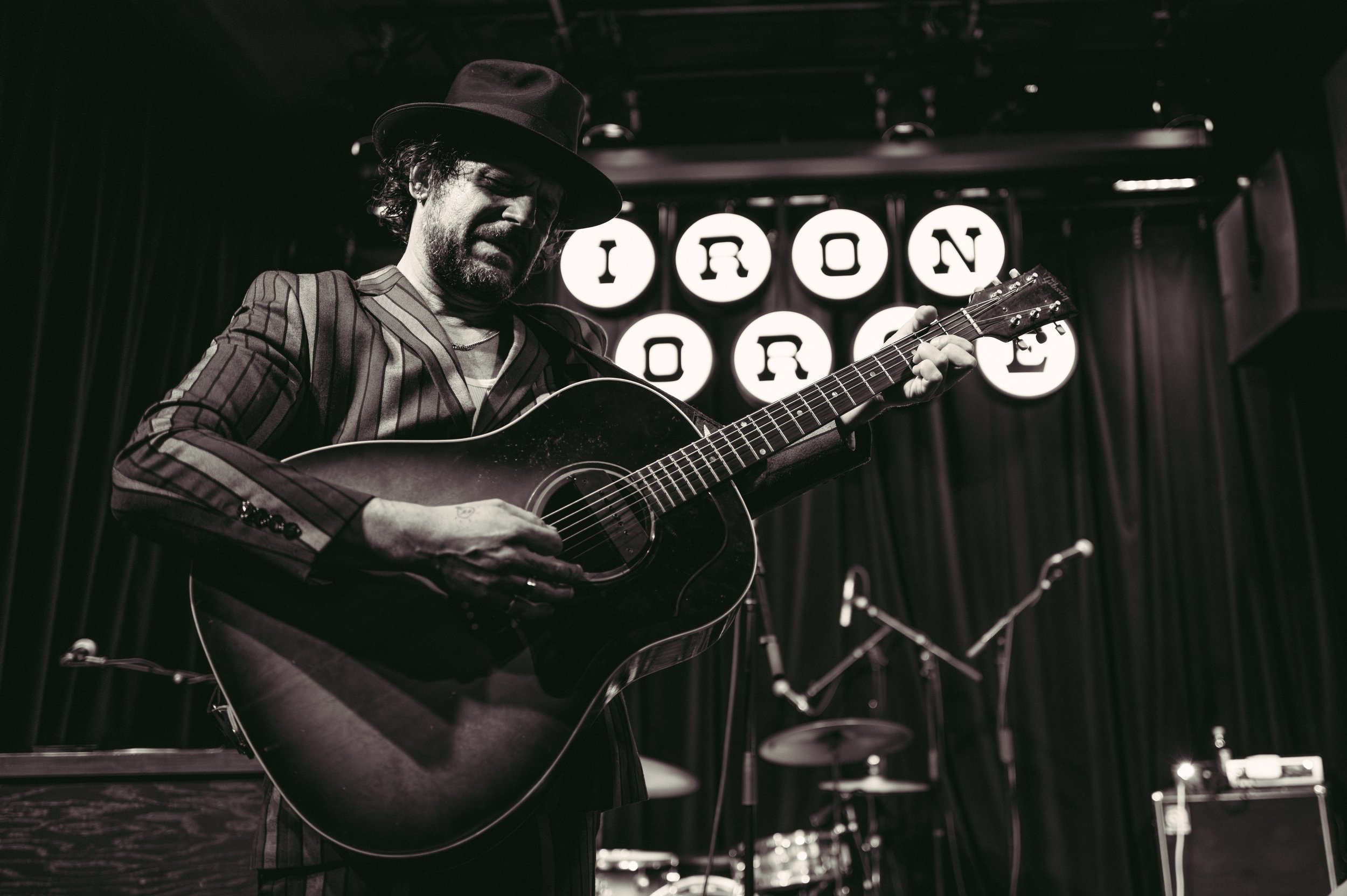 A man with a hat playing an acoustic guitar on a stage with illuminated letters spelling 'IRON HORSE' in the background. Langhorne Slim.